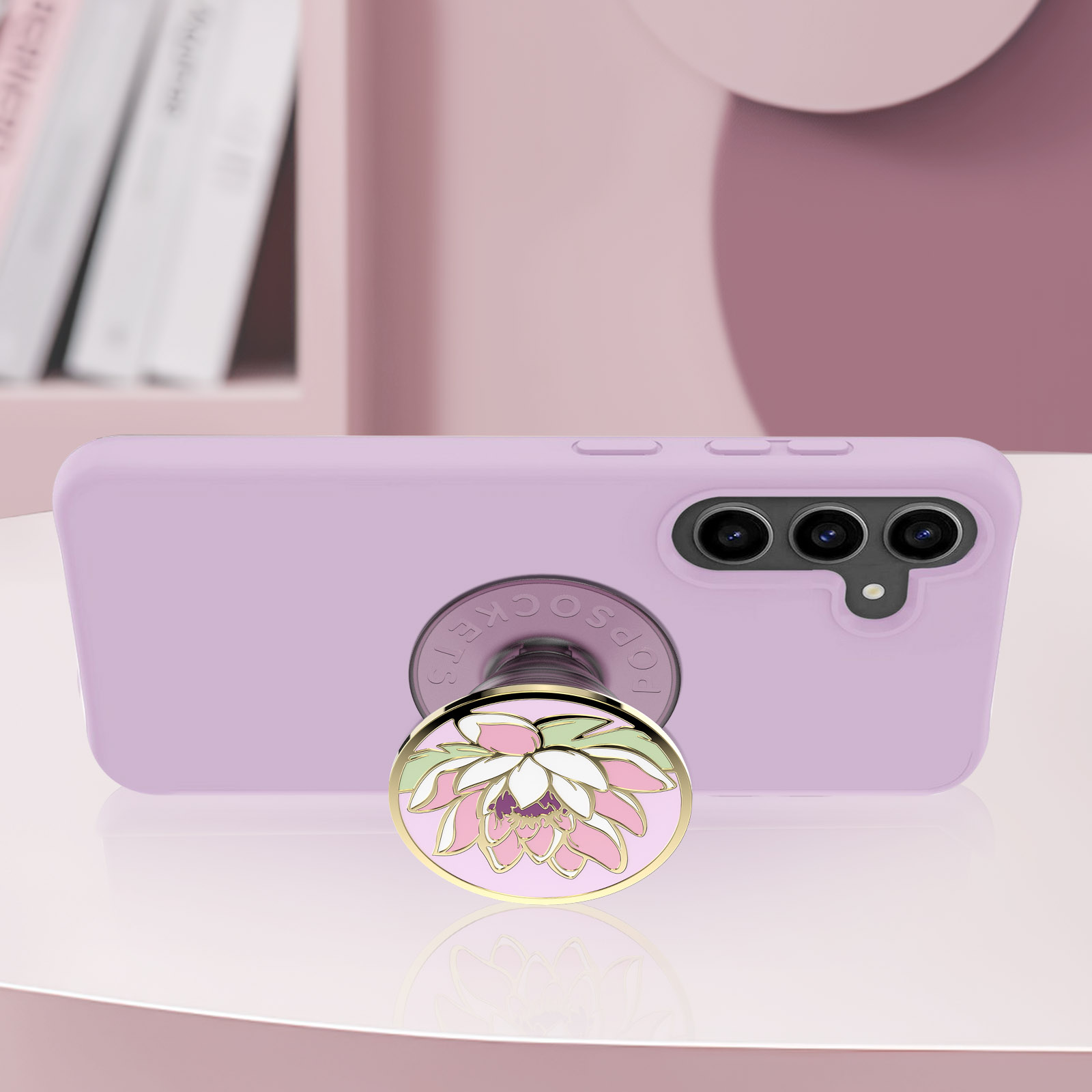 Un telefono lilla con un PopSocket a forma di fiore di loto. Lo sfondo presenta una mensola con libri e una superficie rosa.