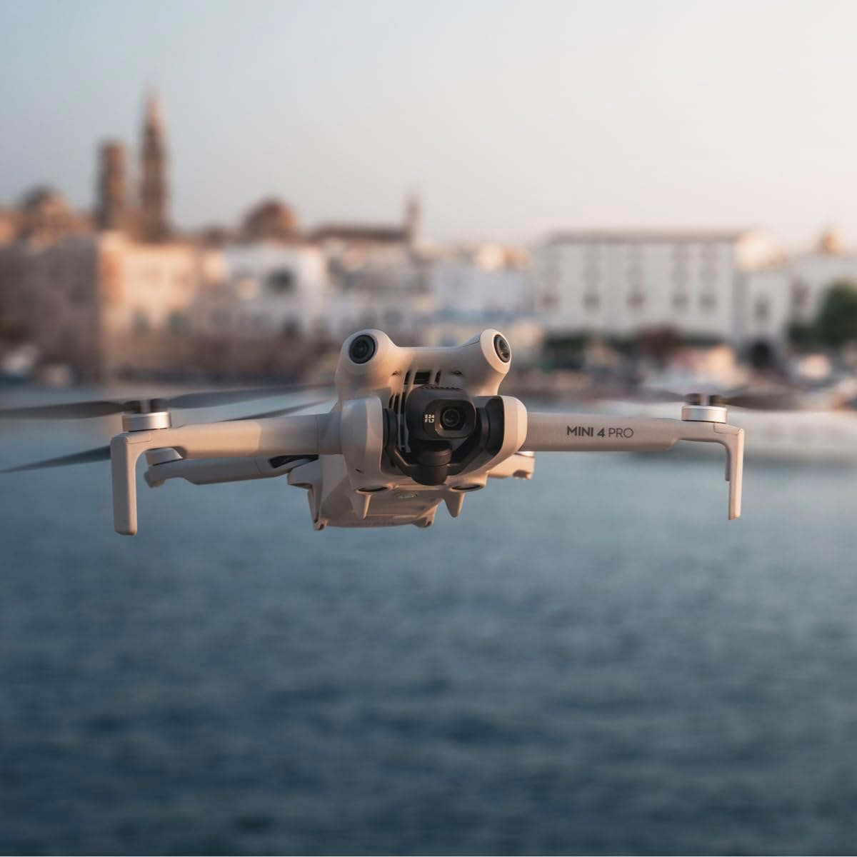 Eine DJI Mini 4 Pro Drohne schwebt über Wasser, mit einer verschwommenen Küstenstadt im Hintergrund. Die Drohne ist hellgrau und hat eine Kamera.