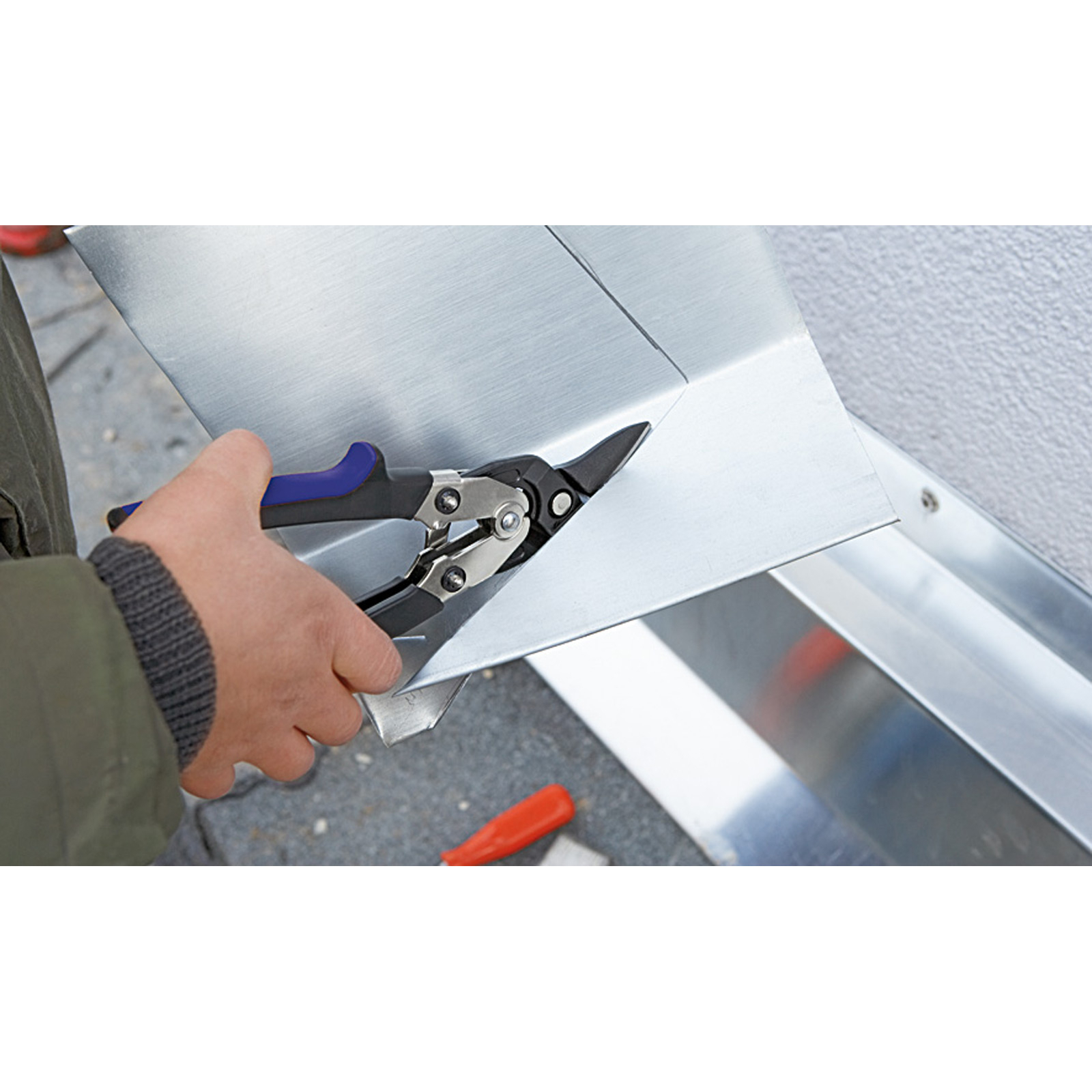 Osoba używa nożyc do blachy do cięcia metalowej blachy. Blacha przylega do ściany. Widoczne jest czerwone narzędzie.