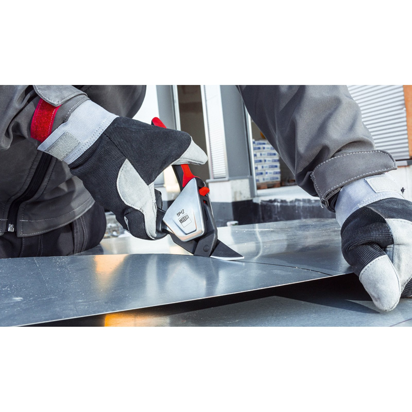 Ein Arbeiter schneidet Metall mit einem Werkzeug. Der Arbeiter trägt Handschuhe und graue Arbeitskleidung. Das Werkzeug ist schwarz und silber mit rotem Griff.