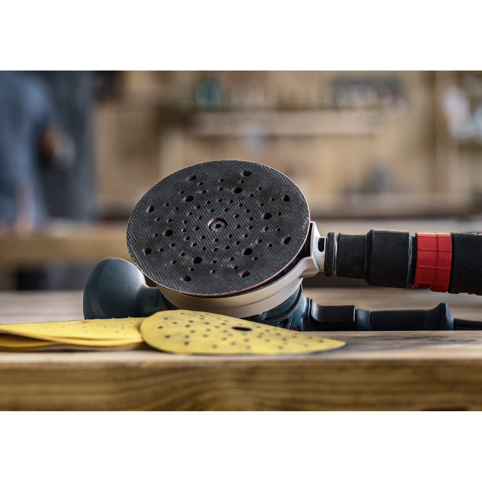 Una levigatrice con tampone nero e carta vetrata gialla su una superficie di legno. Sfondo dell'officina visibile.