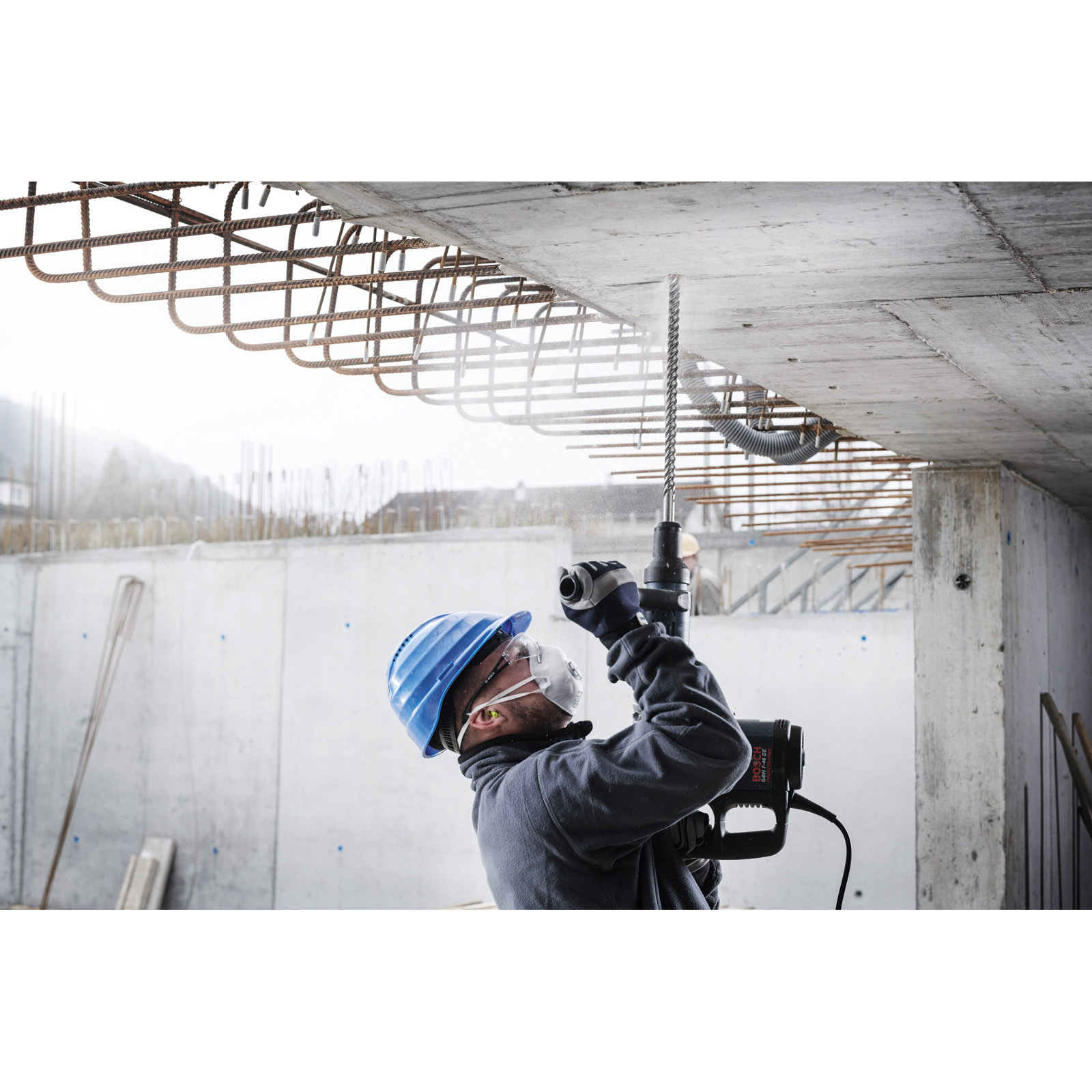 Robotnik budowlany wierci w betonowym suficie wiertarką. Ma niebieski kask, maskę i zatyczki do uszu. Widoczne zbrojenie.