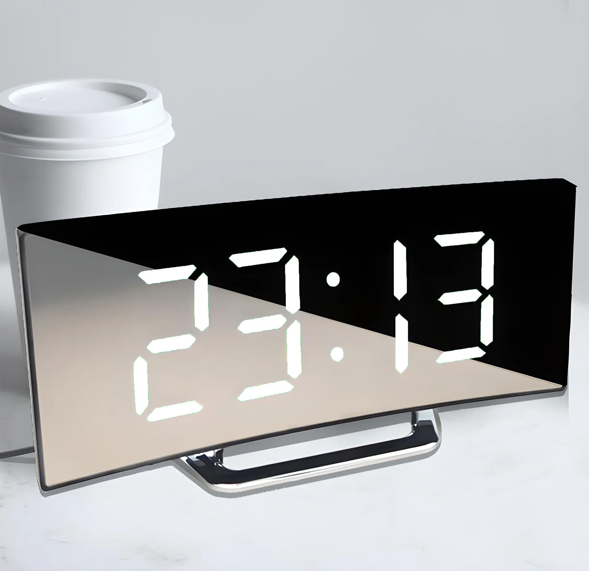 Ein digitaler Wecker, der 23:13 anzeigt, mit einem weißen Kaffeebecher daneben auf einer weißen Oberfläche.