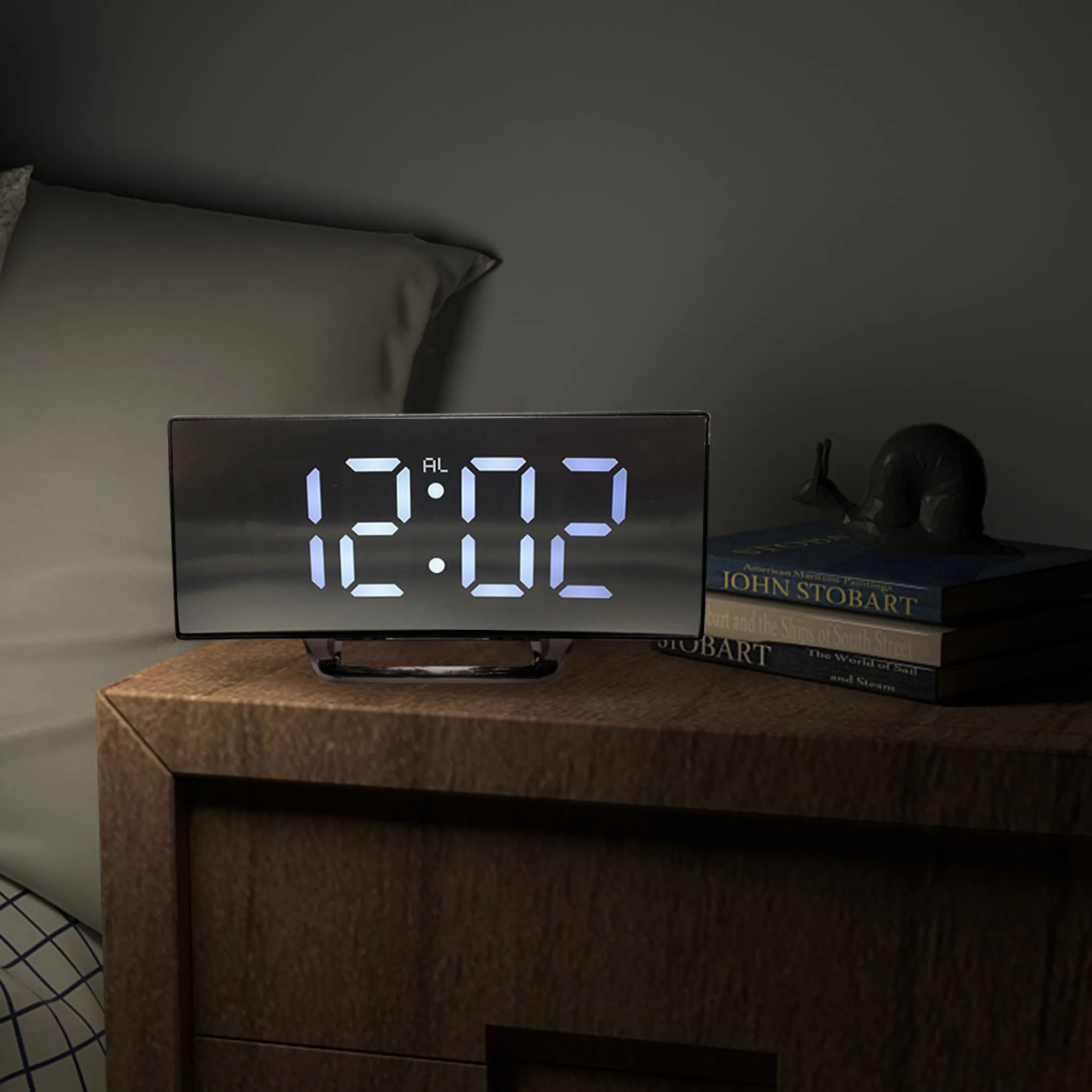 Eine beleuchtete Digitaluhr zeigt 12:02 an. Sie steht auf einem Nachttisch aus Holz mit Büchern und einer Schneckenfigur. Im Hintergrund ist ein Bett.