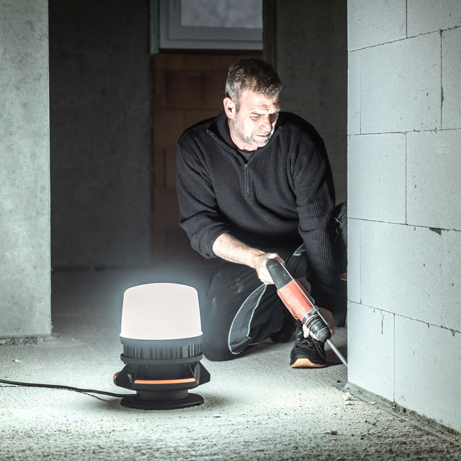 Ein Mann benutzt einen Bohrer in der Nähe einer Wand, beleuchtet von einer Lampe. Die Szene ist auf einer Baustelle.