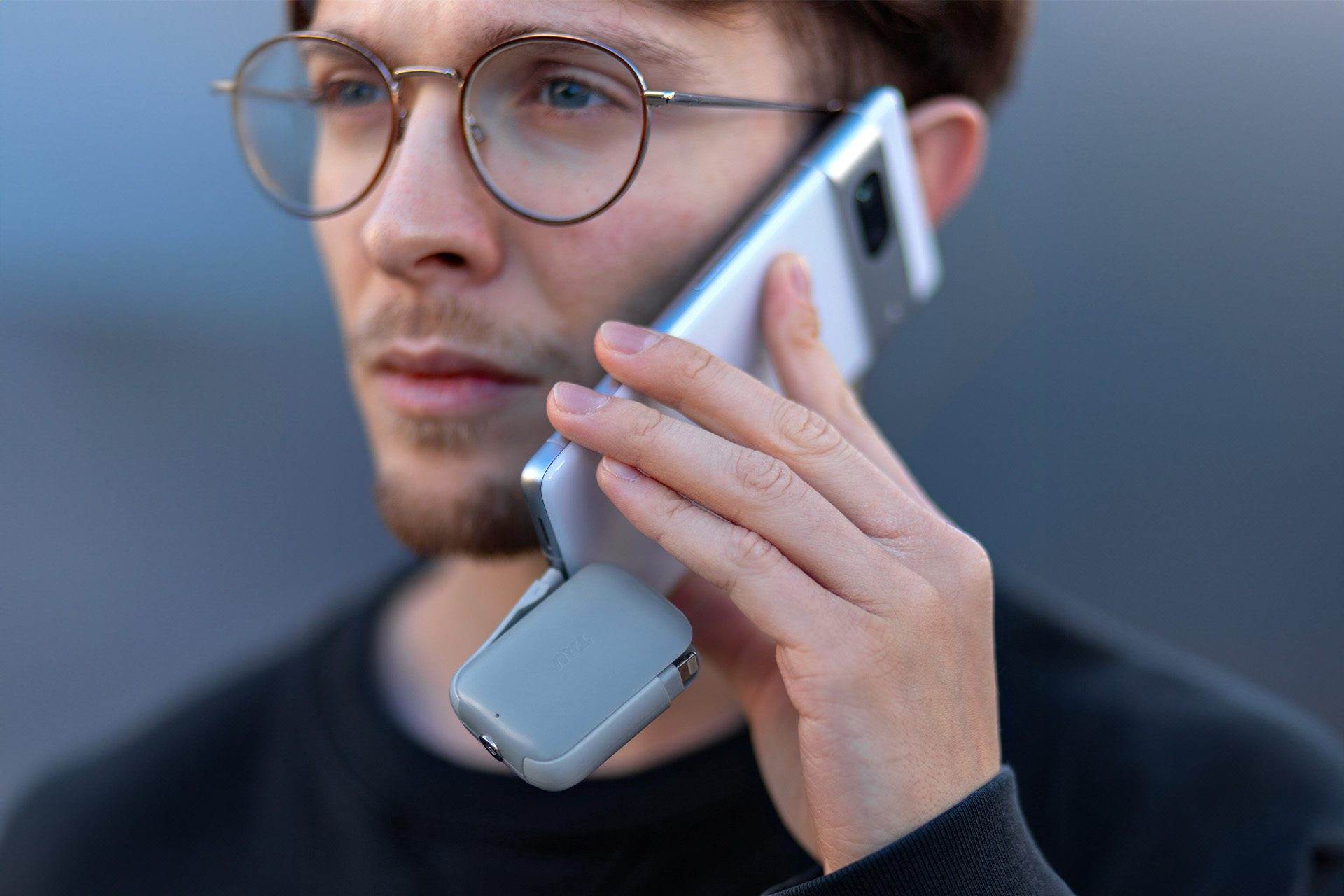 Een man met een bril houdt een telefoon tegen zijn oor. Er is een klein grijs apparaat bevestigd. Hij draagt een zwart shirt.