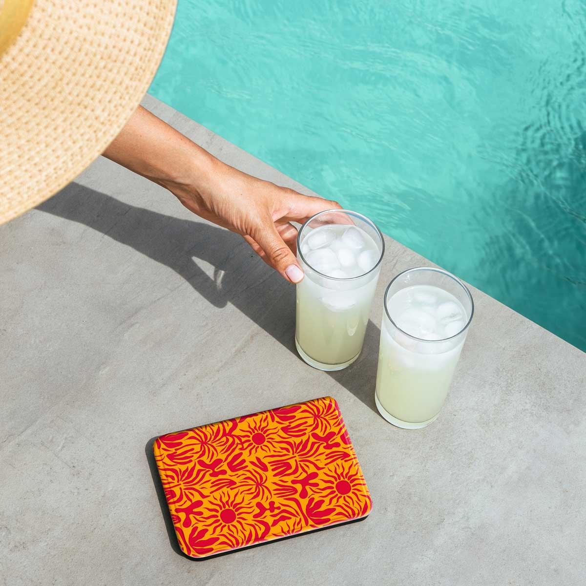 Una mano che afferra un bicchiere di limonata con ghiaccio, accanto a un altro bicchiere e a un sottobicchiere fantasia, vicino a una piscina.