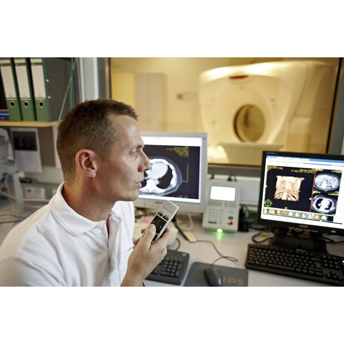 Ein Mann im weißen Hemd hält ein Gerät. Monitore zeigen medizinische Scans. Im Hintergrund befindet sich ein CT-Scanner.