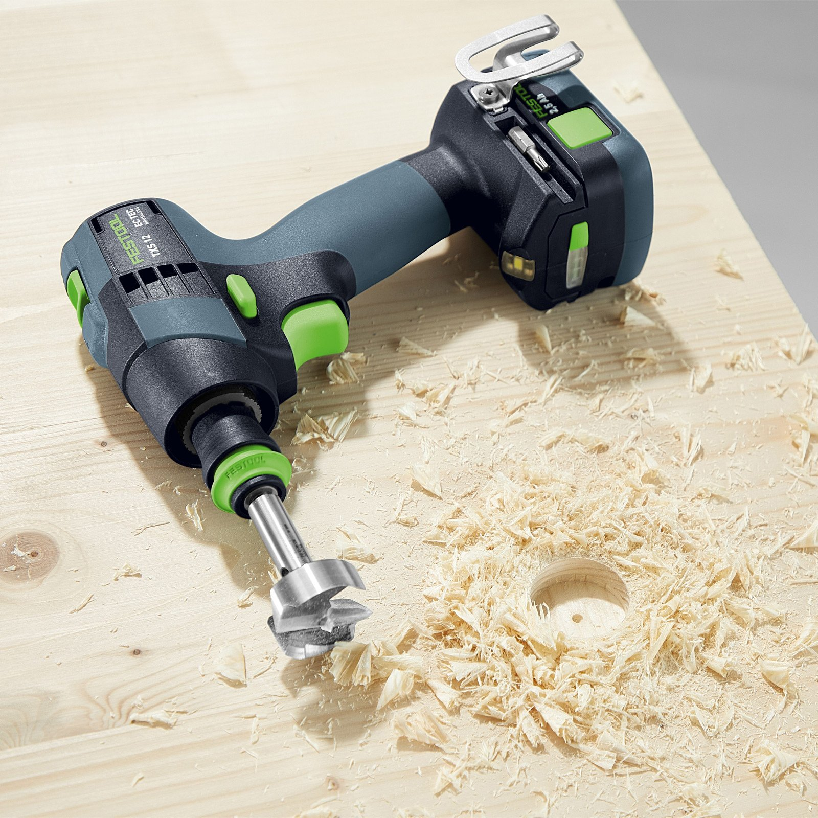 Un taladro inalámbrico perfora un agujero en madera. Virutas de madera rodean el agujero. El taladro es negro, verde y gris.