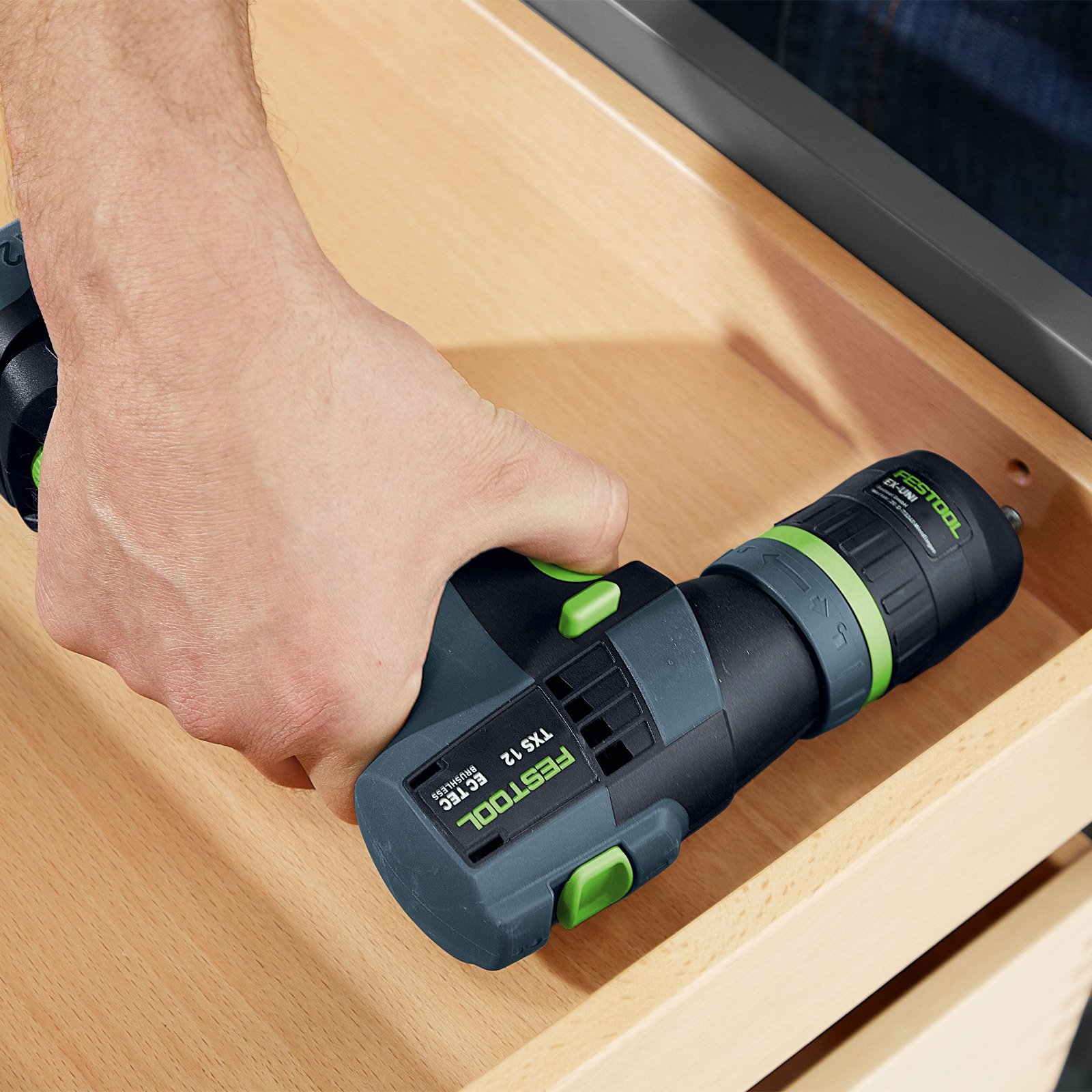 Mano sosteniendo un taladro Festool TXS 12, colocado en un cajón de madera. El taladro es negro y verde.