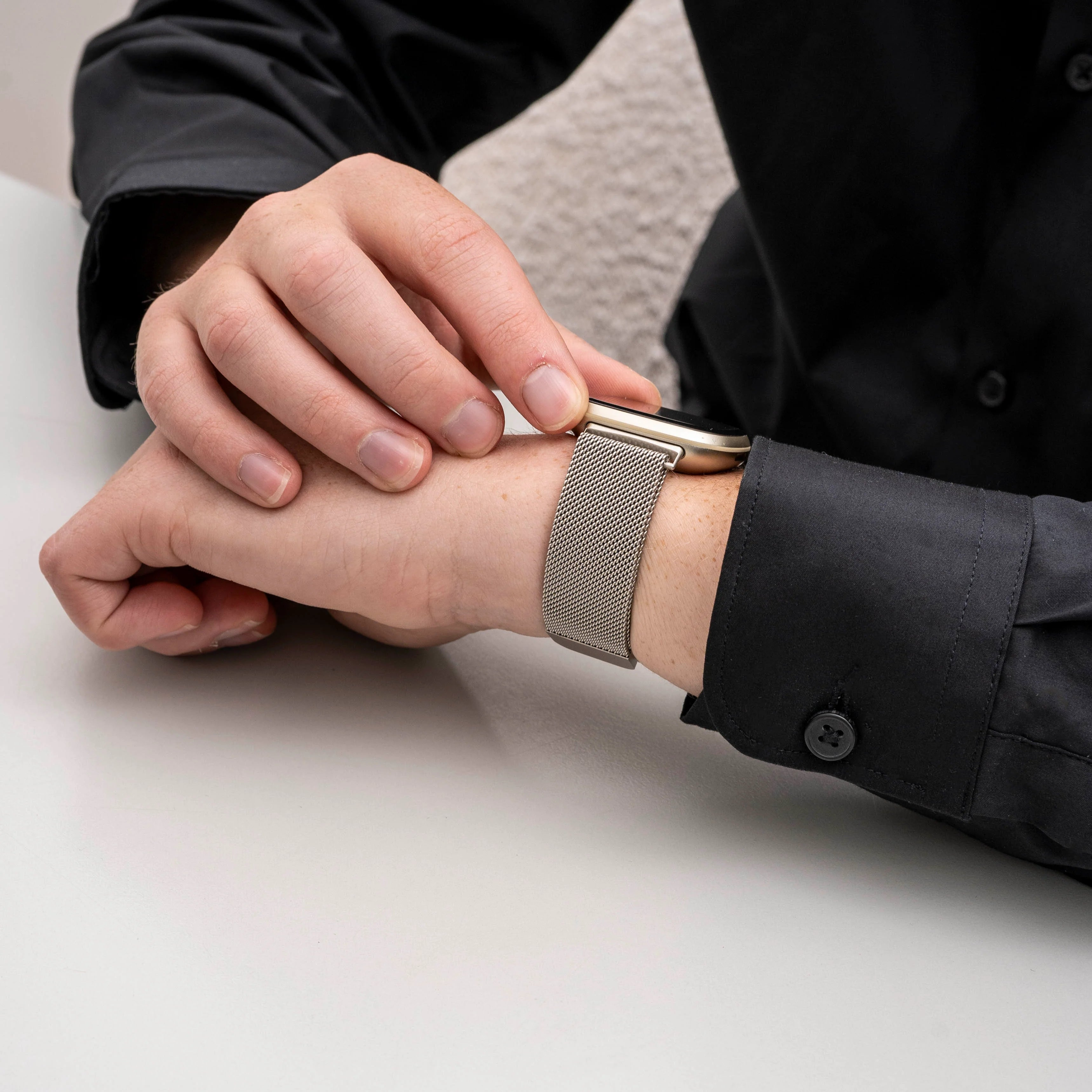 Eine Person in einem schwarzen Hemd justiert eine Smartwatch mit silbernem Mesh-Armband. Die Uhr ist an ihrem Handgelenk.