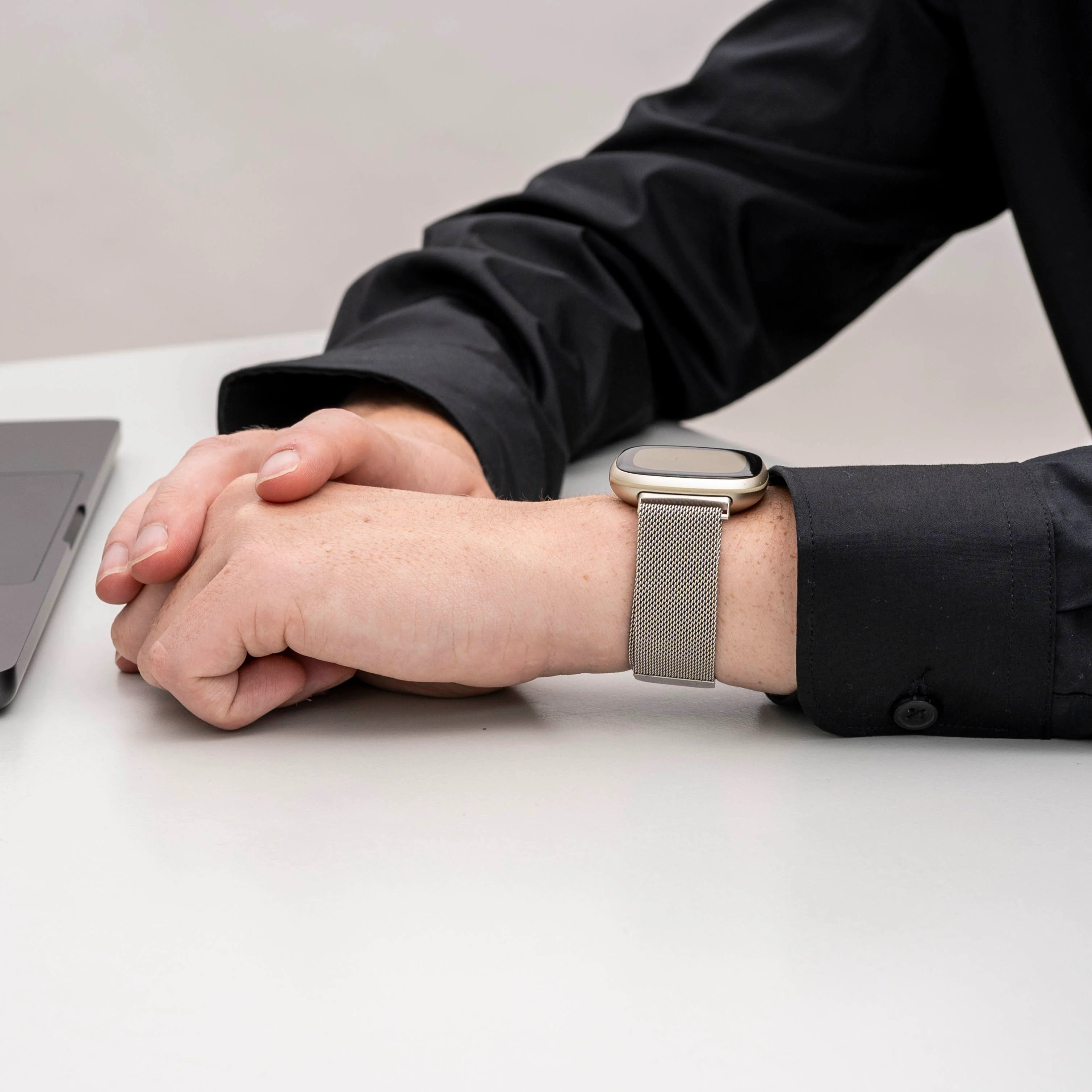 Das Handgelenk einer Person mit einer silbernen Uhr. Die Person trägt ein schwarzes Hemd, im Hintergrund ist ein Laptop zu sehen.