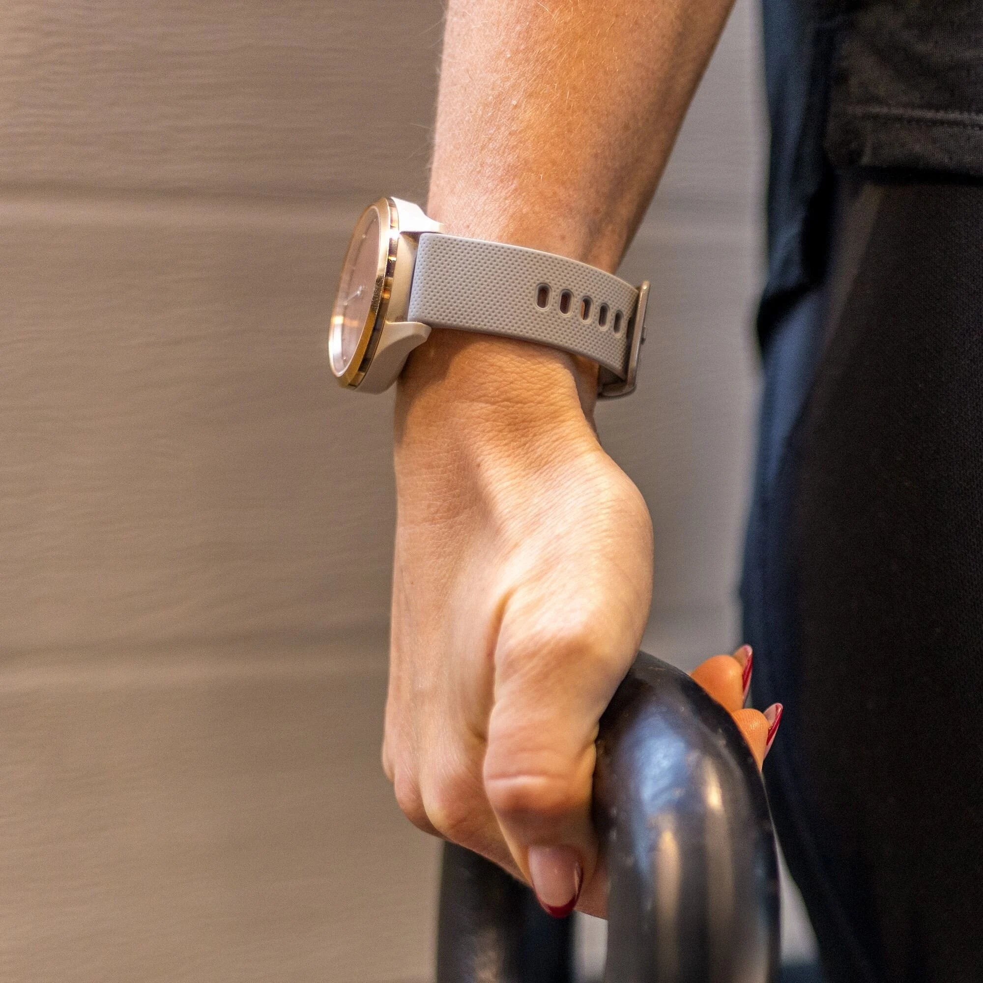 Arm einer Person mit Smartwatch. Die Hand hält eine schwarze Kettlebell. Nägel sind rot lackiert. Der Hintergrund ist eine hellgraue Wand.