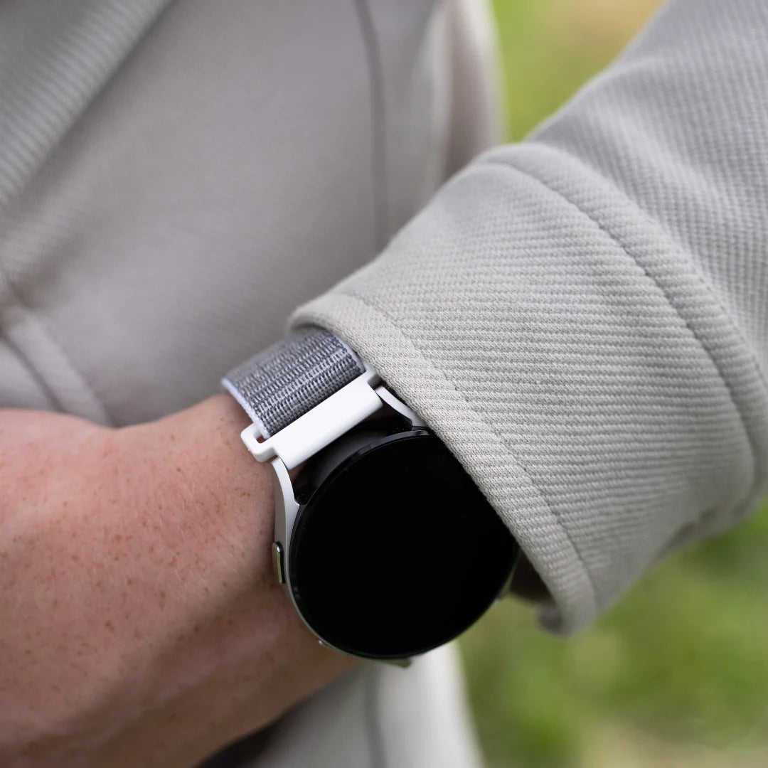 Das Handgelenk einer Person mit einer Smartwatch. Die Uhr hat einen schwarzen Bildschirm und ein graues Stoffband. Der Ärmel eines hellgrauen Mantels ist sichtbar.