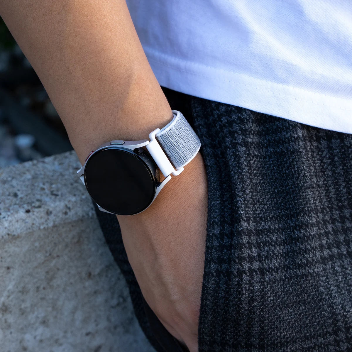 Handgelenk mit Smartwatch. Die Uhr hat einen schwarzen Bildschirm, weißen Rahmen und graues Armband. Die Person trägt ein weißes Hemd und dunkle Hose.