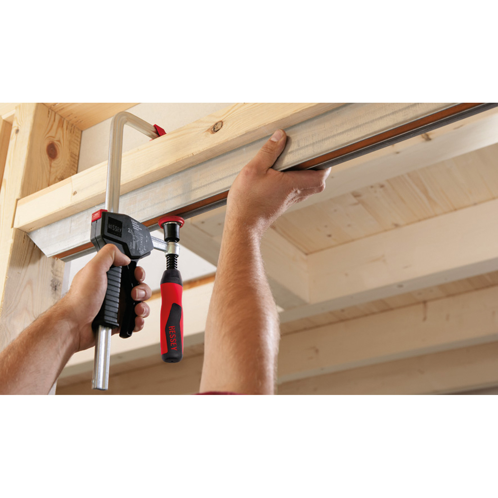 Osoba używa zacisku do utrzymywania drewnianej belki. Zacisk jest czarny i czerwony, a belka jasnobrązowa.