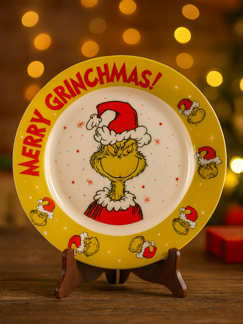 Auf einem Holzständer steht eine Grinchmas-Platte. Sie zeigt den Grinch mit einem Weihnachtsmannhut. Der Hintergrund hat Bokeh-Lichter und einen Weihnachtsbaum.