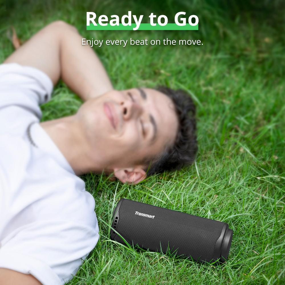Mężczyzna relaksuje się na trawie z czarnym głośnikiem Tronsmart. Tekst brzmi 'Ready to Go' i 'Enjoy every beat on the move'.