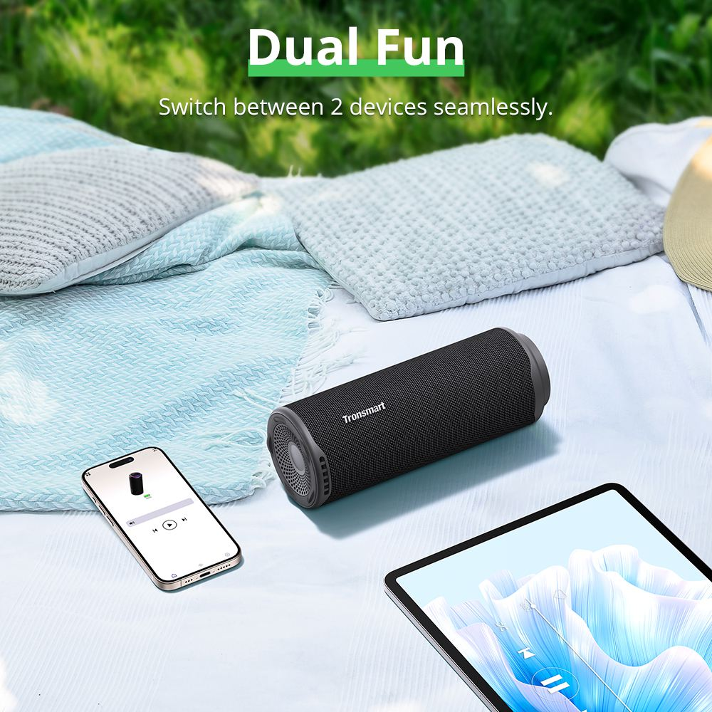 Czarny głośnik Tronsmart, telefon, tablet i poduszki na kocu umieszczone na trawie. Fraza "Dual Fun" znajduje się na górze.