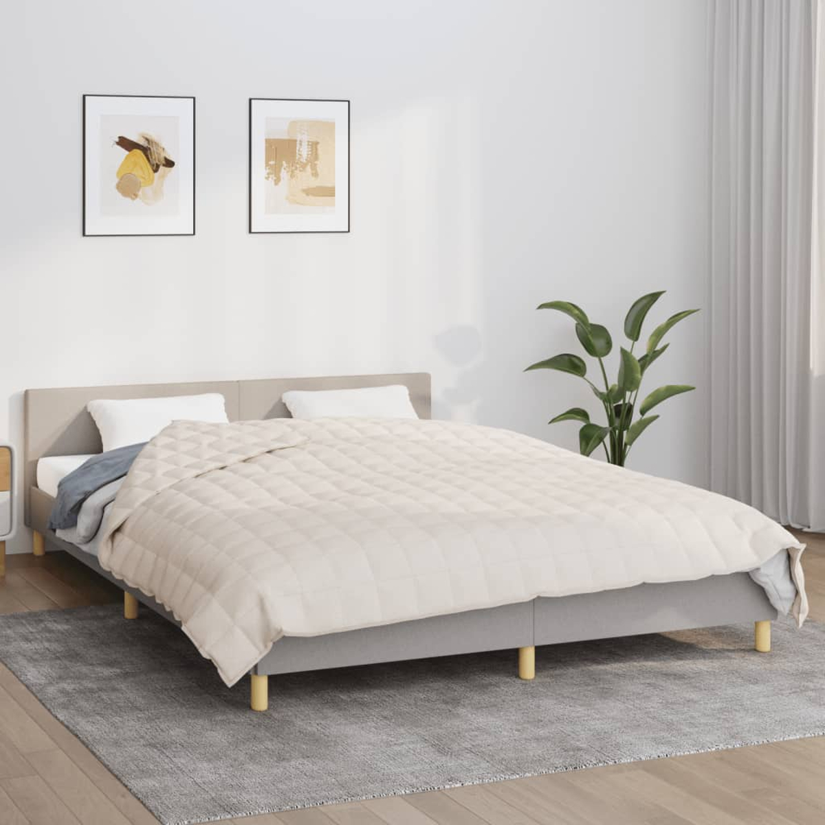 Ein Bett mit einer beigen Bettdecke. Zwei gerahmte Kunstwerke an der Wand. Eine Topfpflanze rechts. Ein grauer Teppich auf dem Boden.