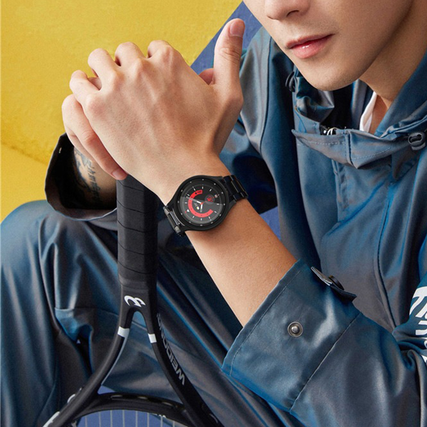 Ein Mann trägt eine schwarze Uhr und hält einen Tennisschläger. Er trägt eine blaue Jacke. Der Hintergrund ist gelb.
