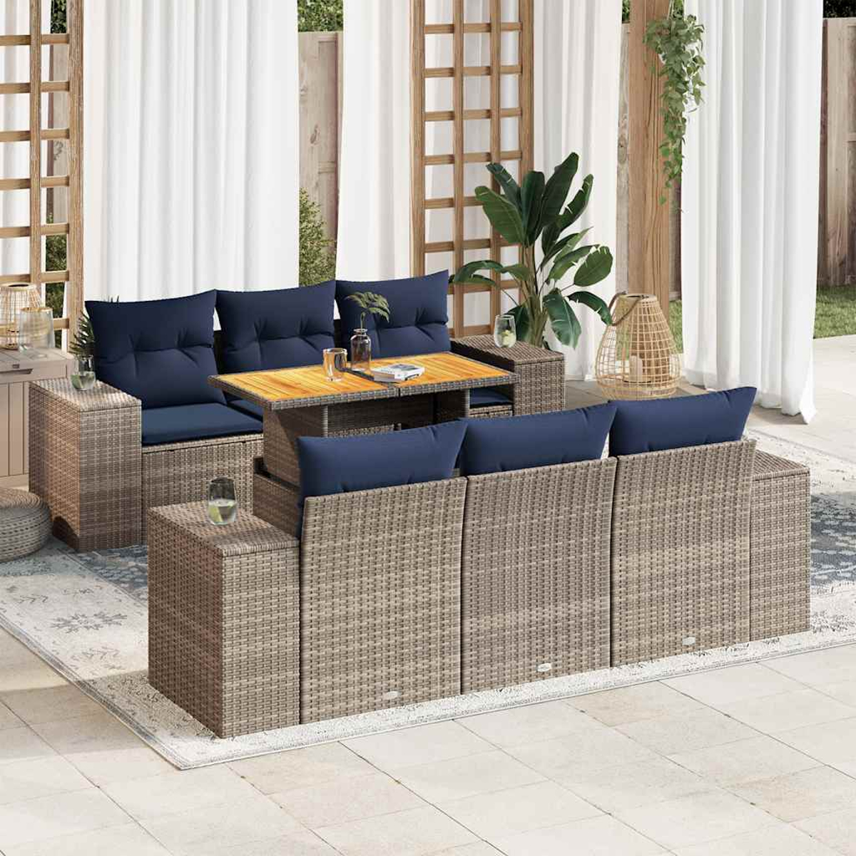 Gartenmöbel-Set aus Korbgeflecht mit blauen Kissen, einem Holztisch und weißen Vorhängen im Hintergrund. Auf dem Boden liegt ein Teppich.