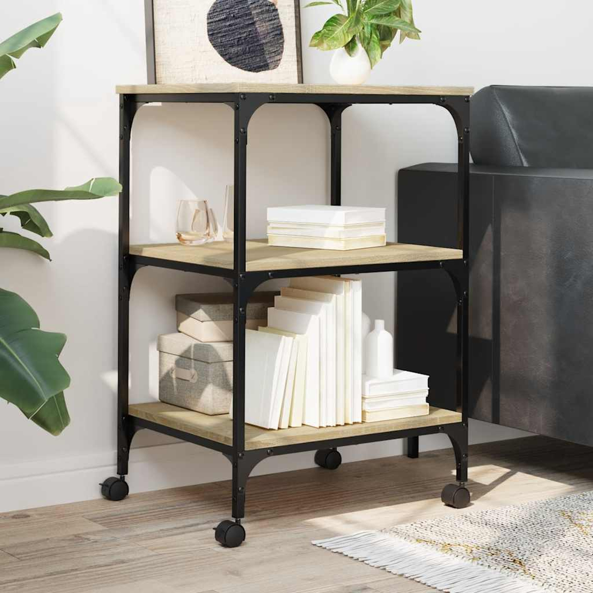 Ein dreistöckiges Regal auf Rädern mit Büchern, Kisten und Dekoration. Der Rahmen ist schwarz, die Regale sind helles Holz.