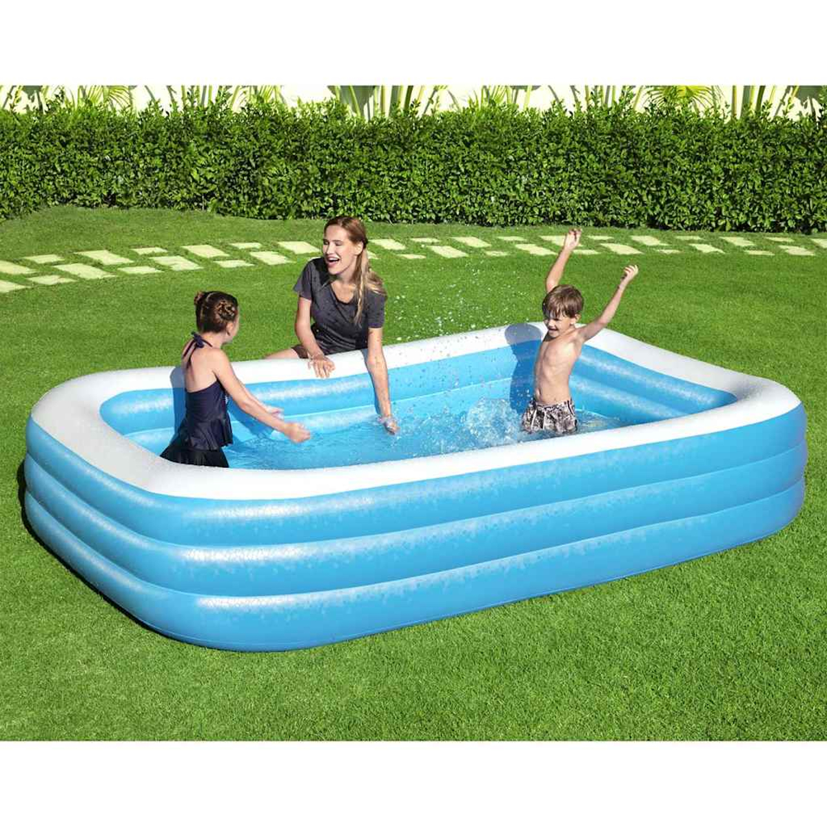 Eine Frau und zwei Kinder spielen in einem blauen Planschbecken auf grünem Gras und spritzen mit Wasser. Im Hintergrund befindet sich eine grüne Hecke.