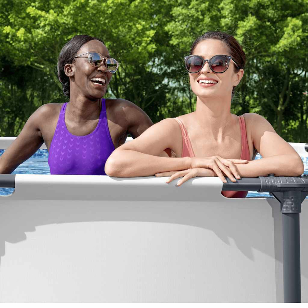 Due donne con occhiali da sole sorridono in piscina. Una indossa un costume viola, l'altra rosa. Alberi verdi sullo sfondo.