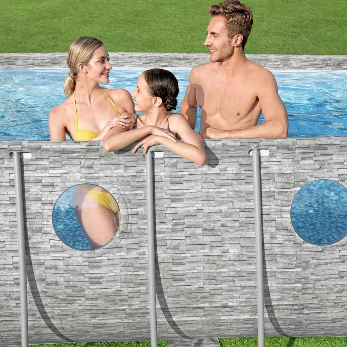 Drei Personen in einem Pool. Eine Frau im gelben Bikini, ein Mädchen in Schwarz und ein Mann. Der Pool hat eine graue Steinmauer.