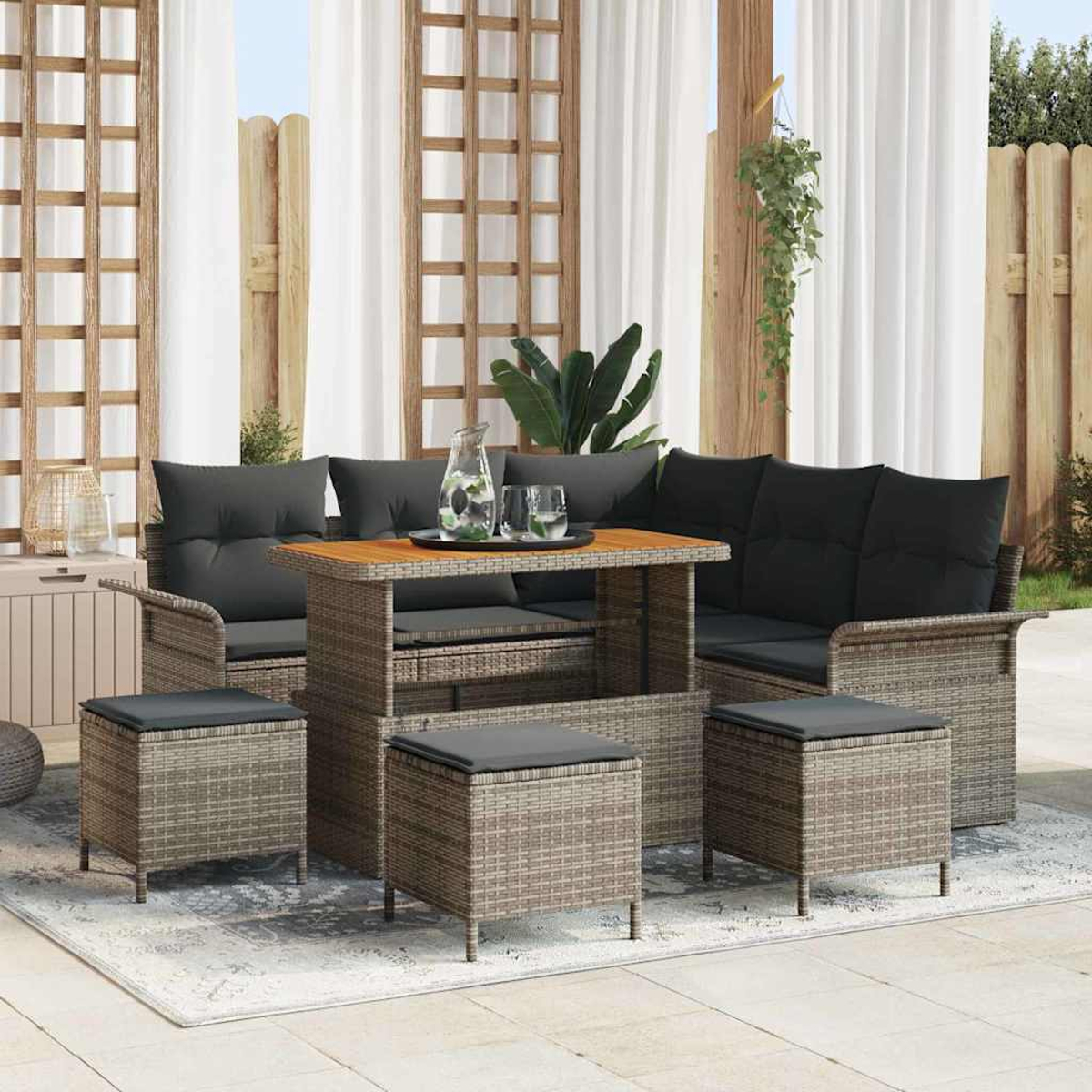 Gartenmöbel-Set aus Rattan mit schwarzen Kissen auf einem Teppich. Tisch mit Gläsern und einer Kanne. Weiße Vorhänge und Holzzaun im Hintergrund.