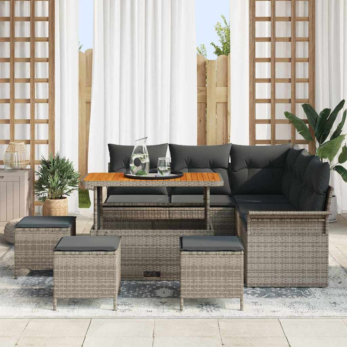 Gartenmöbel-Set. Rattan-Sofa mit grauen Kissen, Holztisch und Hockern. Pflanzen und weiße Vorhänge im Hintergrund.