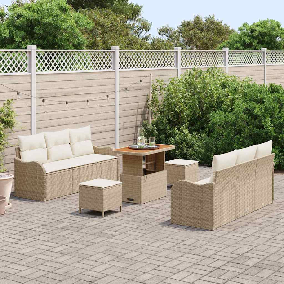 Gartenmöbel-Set mit beigen Sofas, Tisch und Hockern. Auf einer Backstein-Terrasse, mit weißem Zaun und grünen Bäumen im Hintergrund.