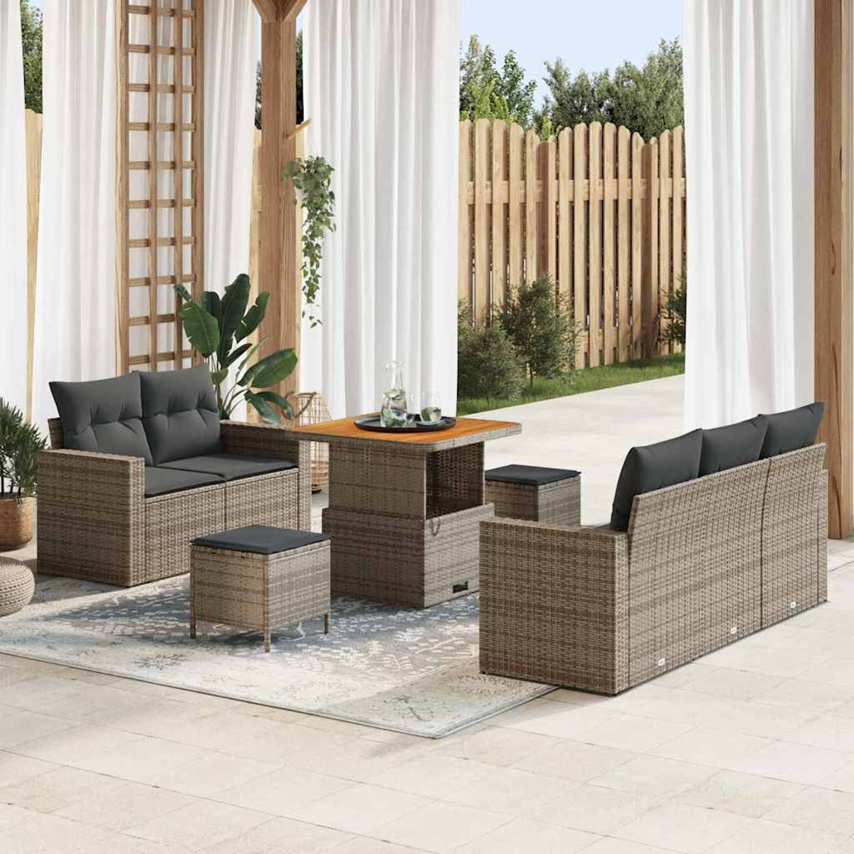 Terrassenset mit gewebten Möbeln, grauen Kissen, Tisch und Hockern. Weiße Vorhänge und Holzzaun im Hintergrund.