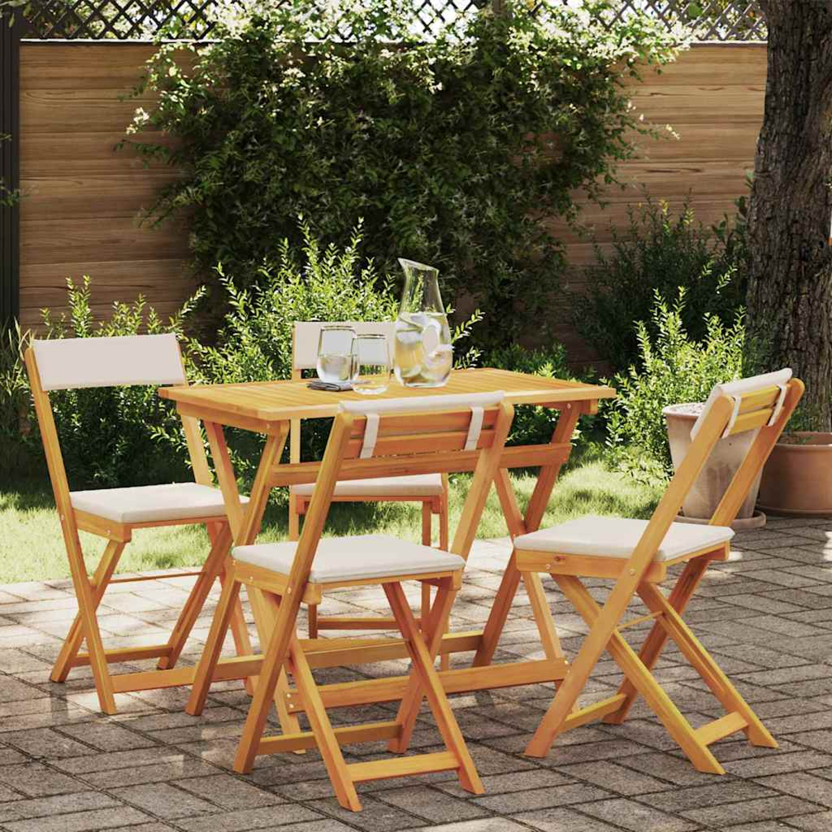 Ein Holztisch und Stühle stehen auf einer Terrasse. Eine Wasserkaraffe und Gläser stehen auf dem Tisch. Dahinter grünes Laub.