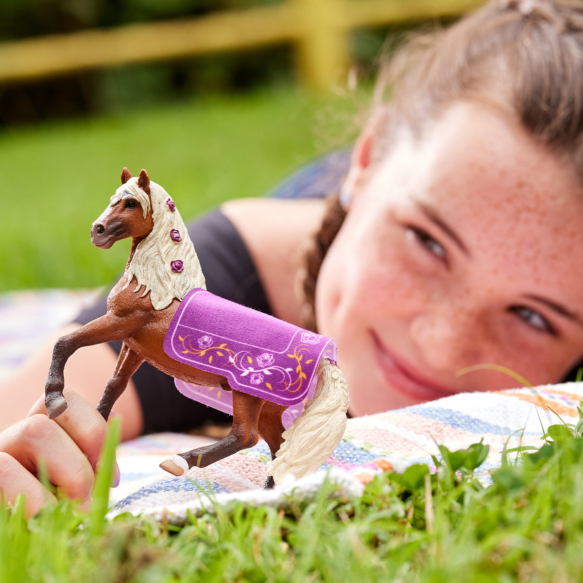 Zabawkowy koń z fioletowym kocem trzymany jest przez rękę. Uśmiechnięta dziewczyna z piegami jest w tle na kocu na trawie.