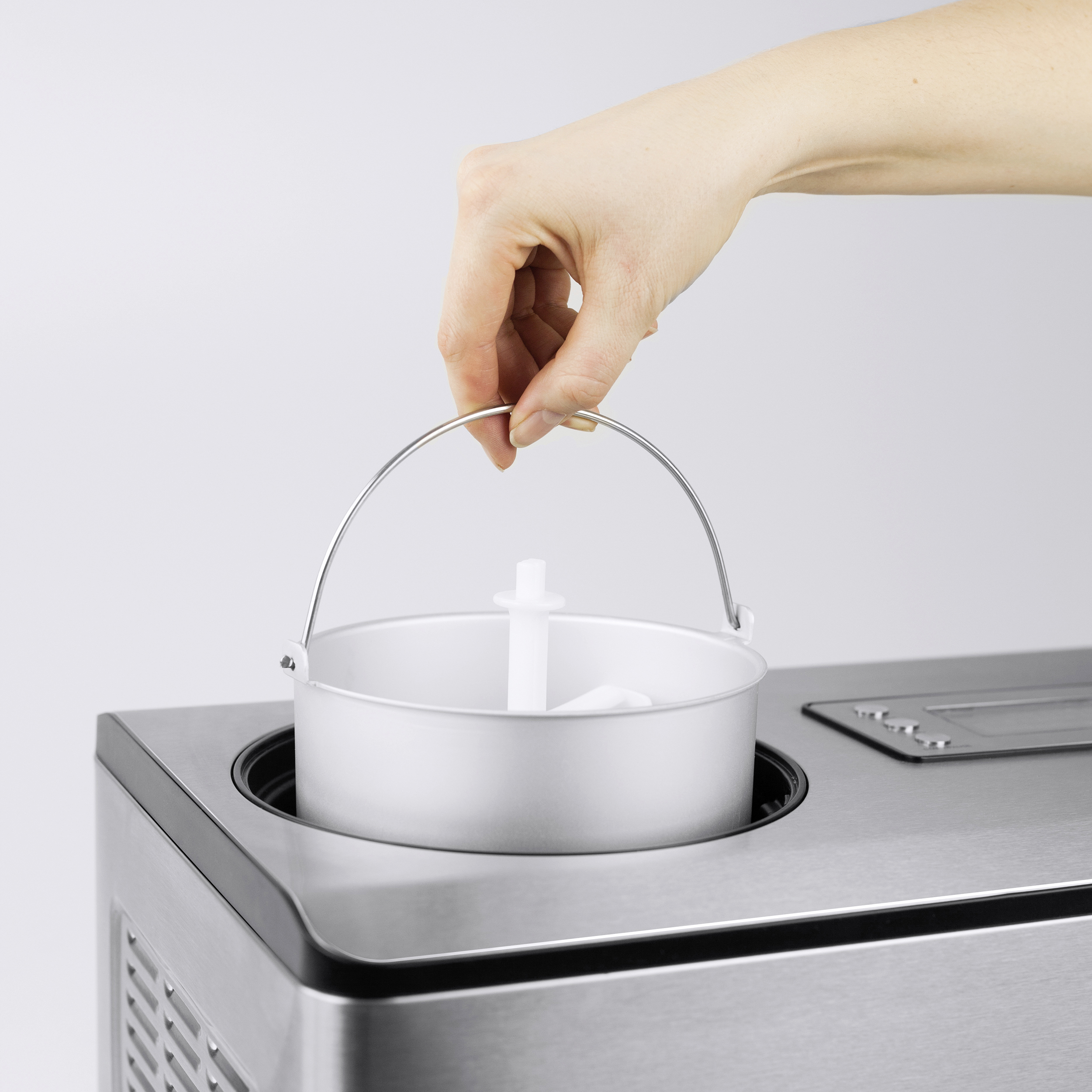 Eine Hand hält den Griff einer Eiscremebehälters. Die Maschine ist silberfarben, der Hintergrund weiß.