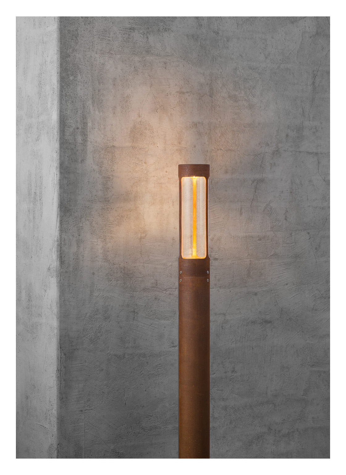 Una lampada cilindrica arrugginita è accesa, posta contro un muro di cemento grigio. La lampada ha una luce gialla brillante all'interno.