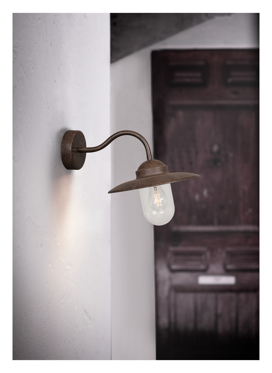 Eine Wandlampe im rustikalen Stil mit gebogenem Arm und klarer Glühbirne. Dahinter ist eine dunkle Holztür zu sehen.