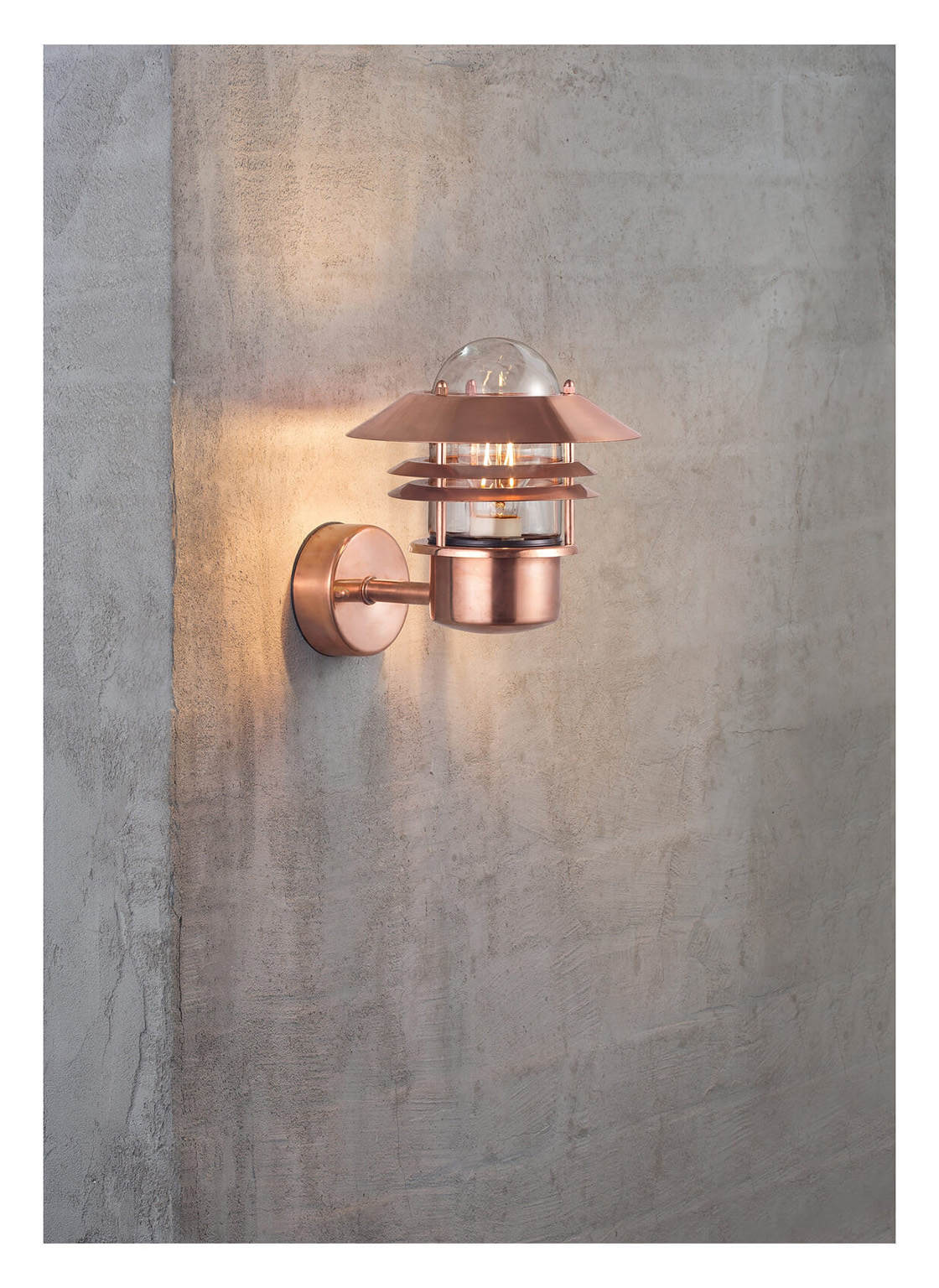 Kupfer-Wandlampe mit Glaskuppel an einer strukturierten grauen Wand. Das Licht ist an und erzeugt ein warmes Leuchten.