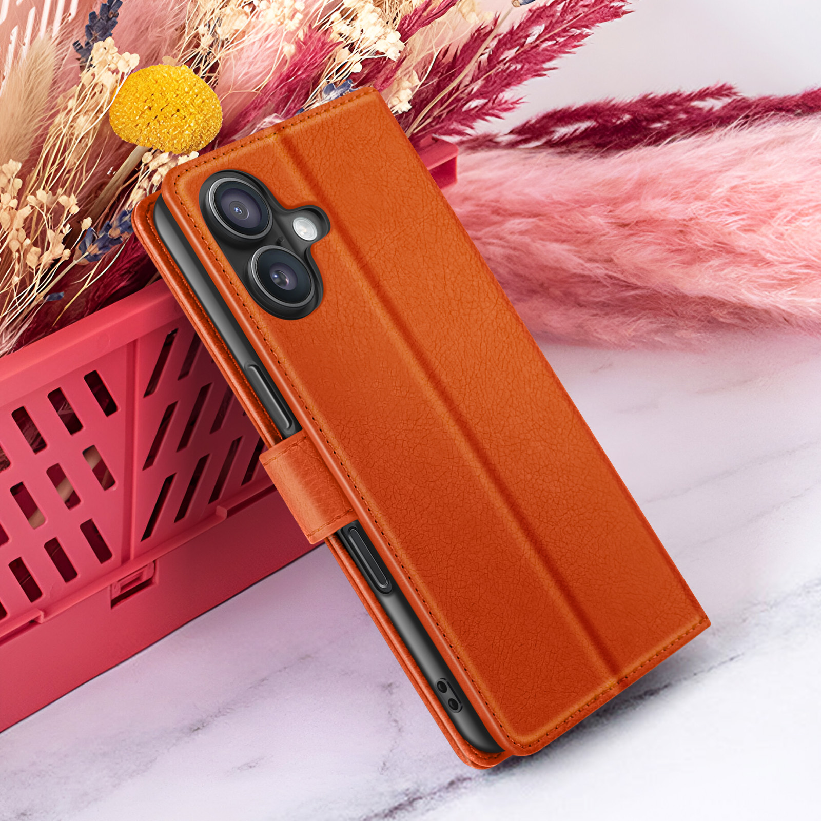 Un étui de téléphone orange repose contre un récipient rouge et des fleurs. L'arrière-plan comporte des fleurs roses et beiges. La surface est en marbre.