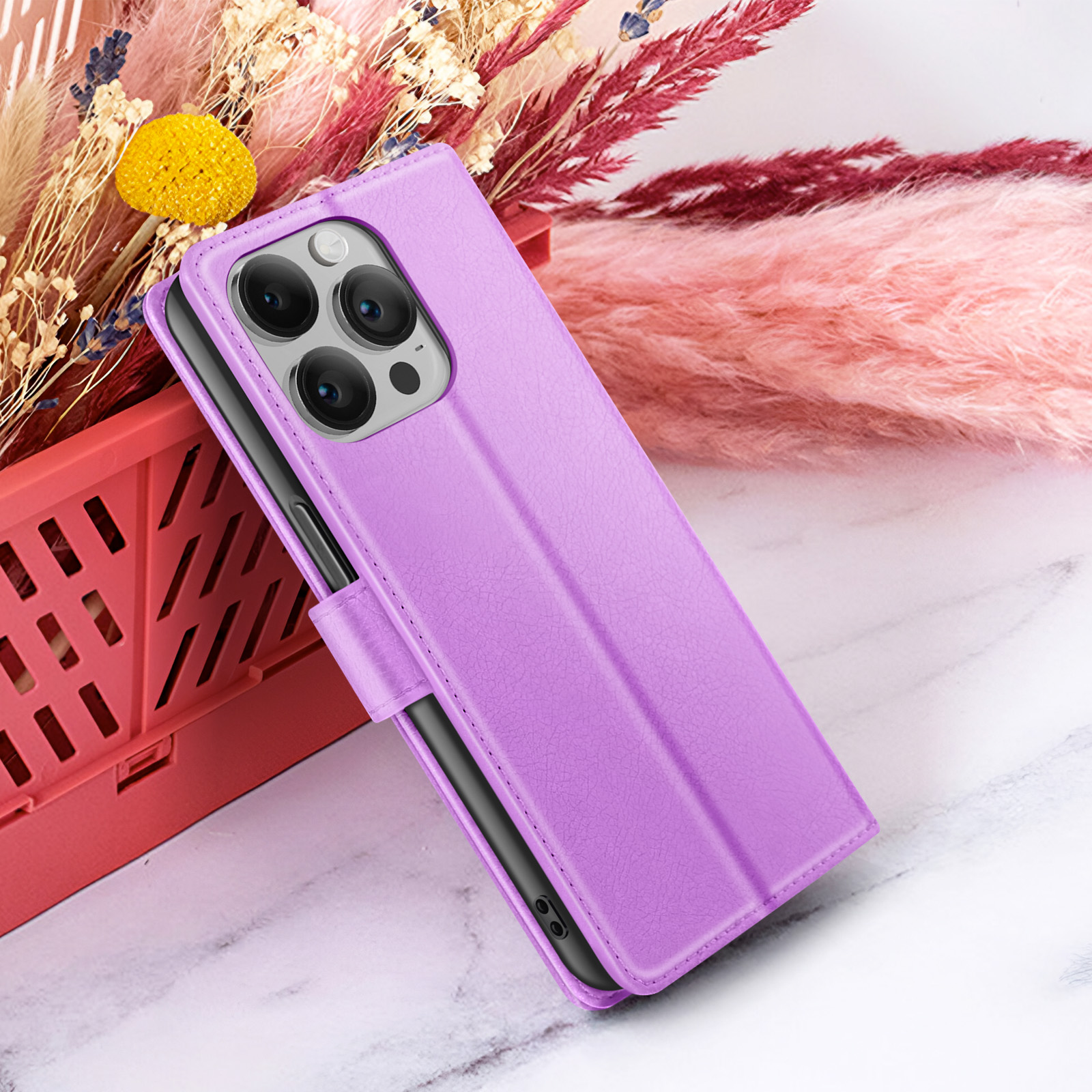 Een paarse telefoonhoes wordt getoond tegen een achtergrond van gedroogde bloemen en een rode container, op een wit marmeren oppervlak.