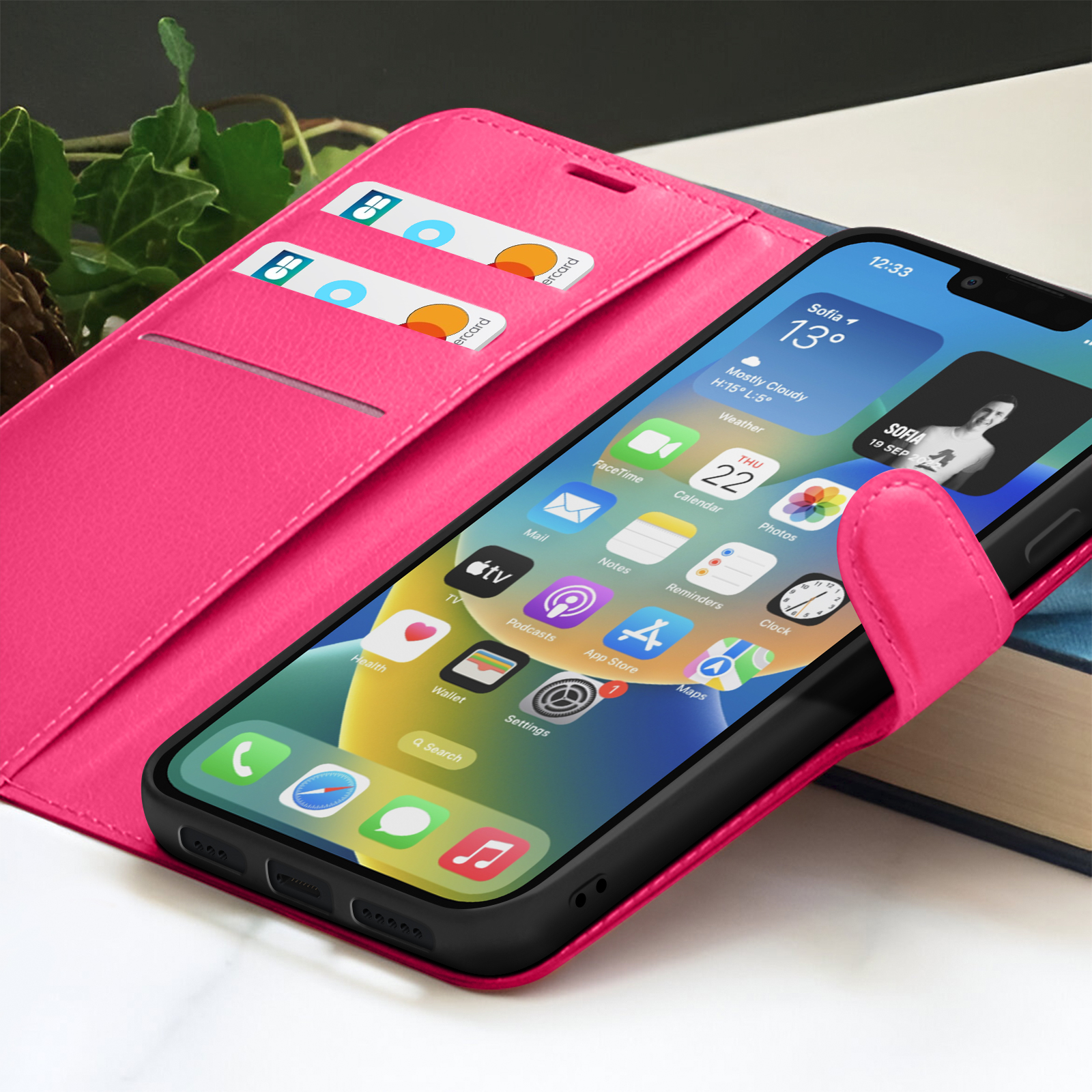 Un smartphone dans un étui rose affiche des icônes d'application. Deux cartes de crédit sont dans l'étui. L'arrière-plan comprend des livres et des feuilles vertes.
