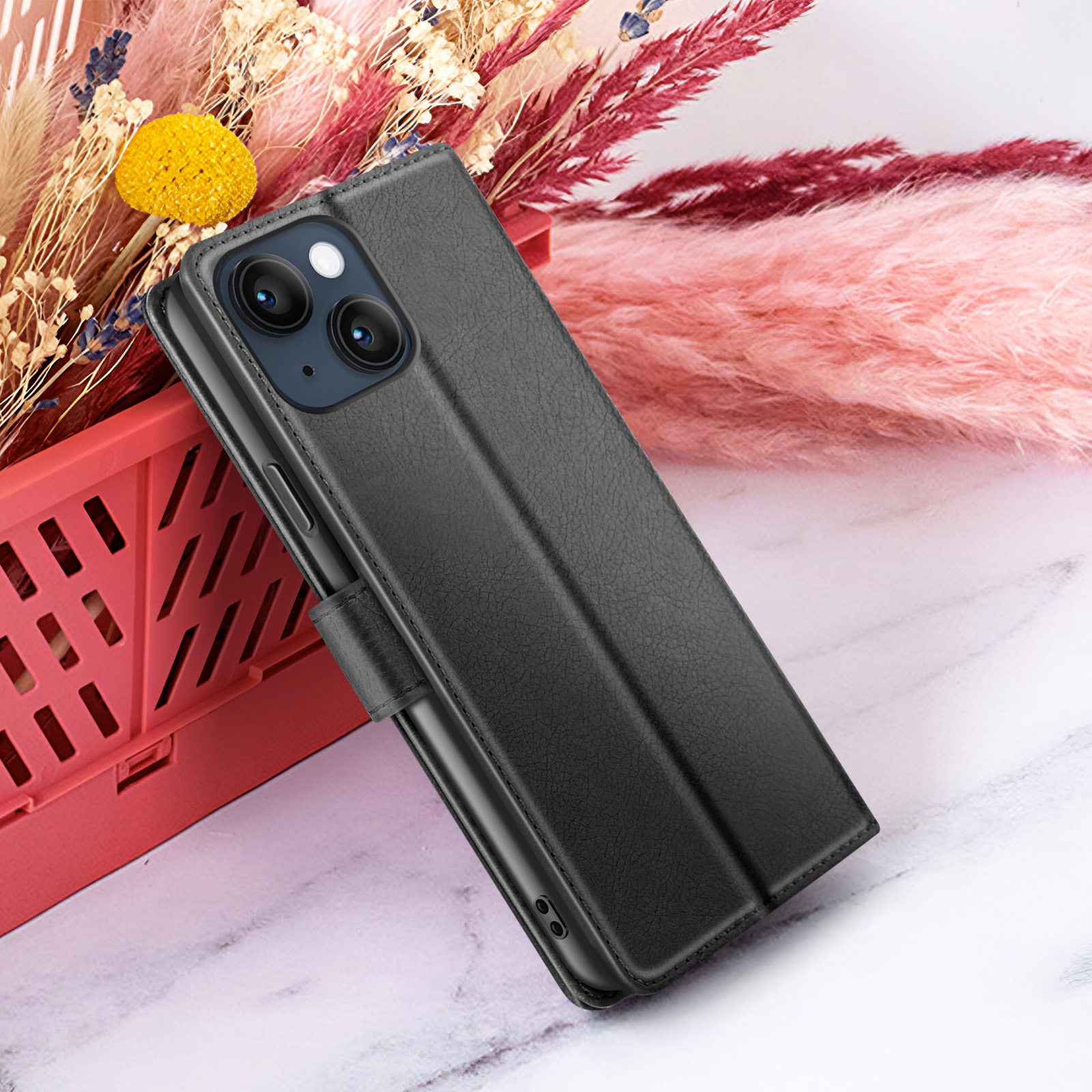 Un étui de téléphone noir est ouvert, révélant un téléphone avec une caméra. Fleurs et un conteneur rouge en arrière-plan.