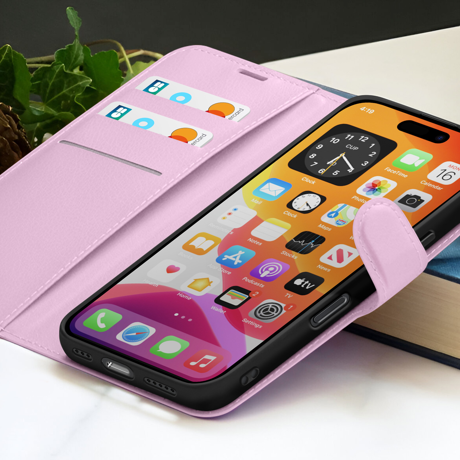 Een smartphone in een roze hoesje toont apps. Er zitten creditcards in de hoes. De achtergrond toont groene bladeren en een boek.