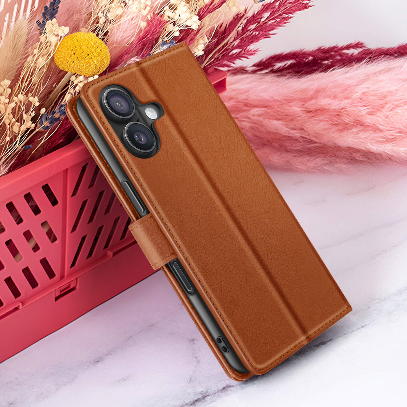 Een bruine telefoonhoes met een telefoon erin staat in de buurt van een rode plastic mand en kleurrijke gedroogde bloemen.