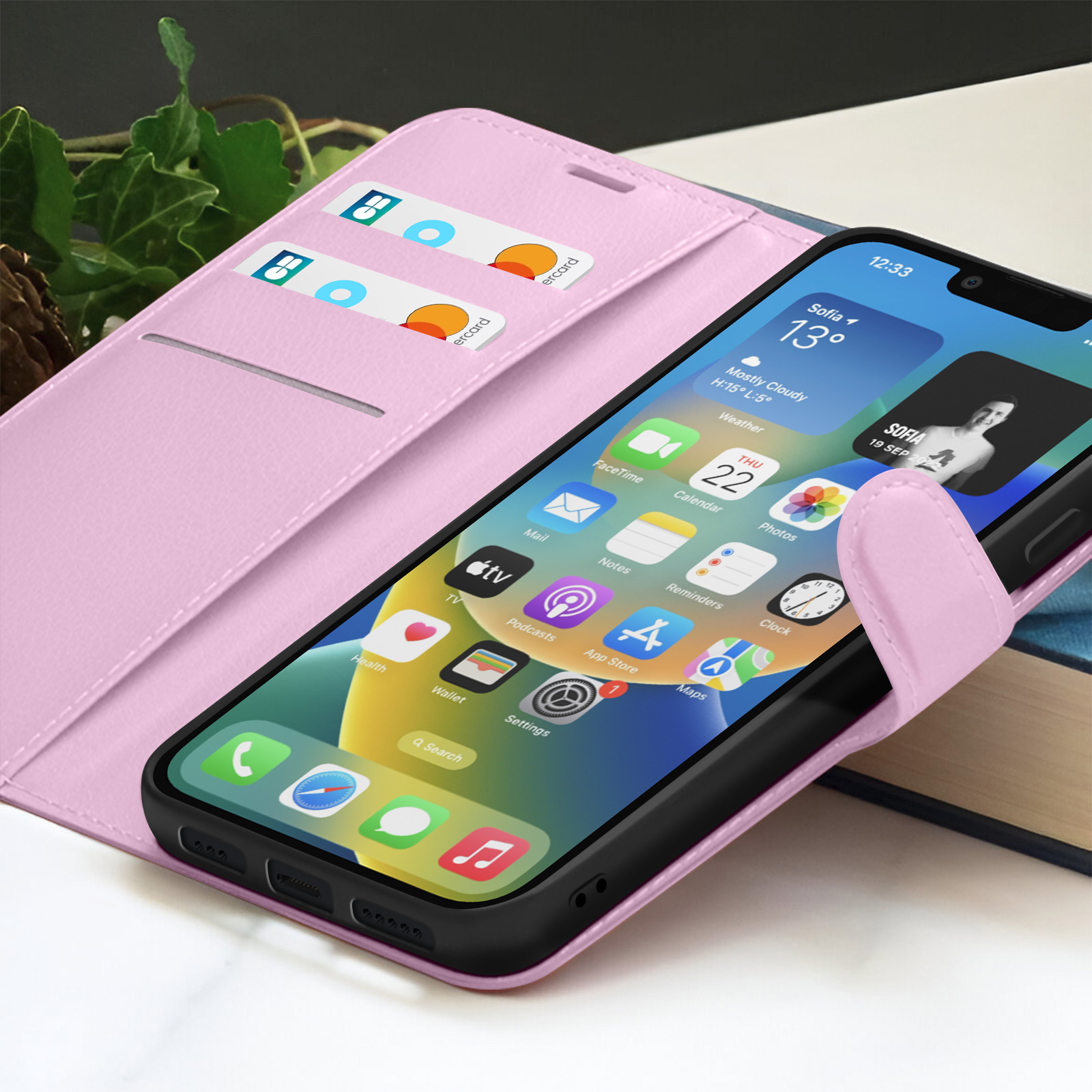 Roze telefoonhoesje met telefoon met app-iconen. Creditcards zichtbaar in de hoes. Achtergrond met bladeren en een boek.