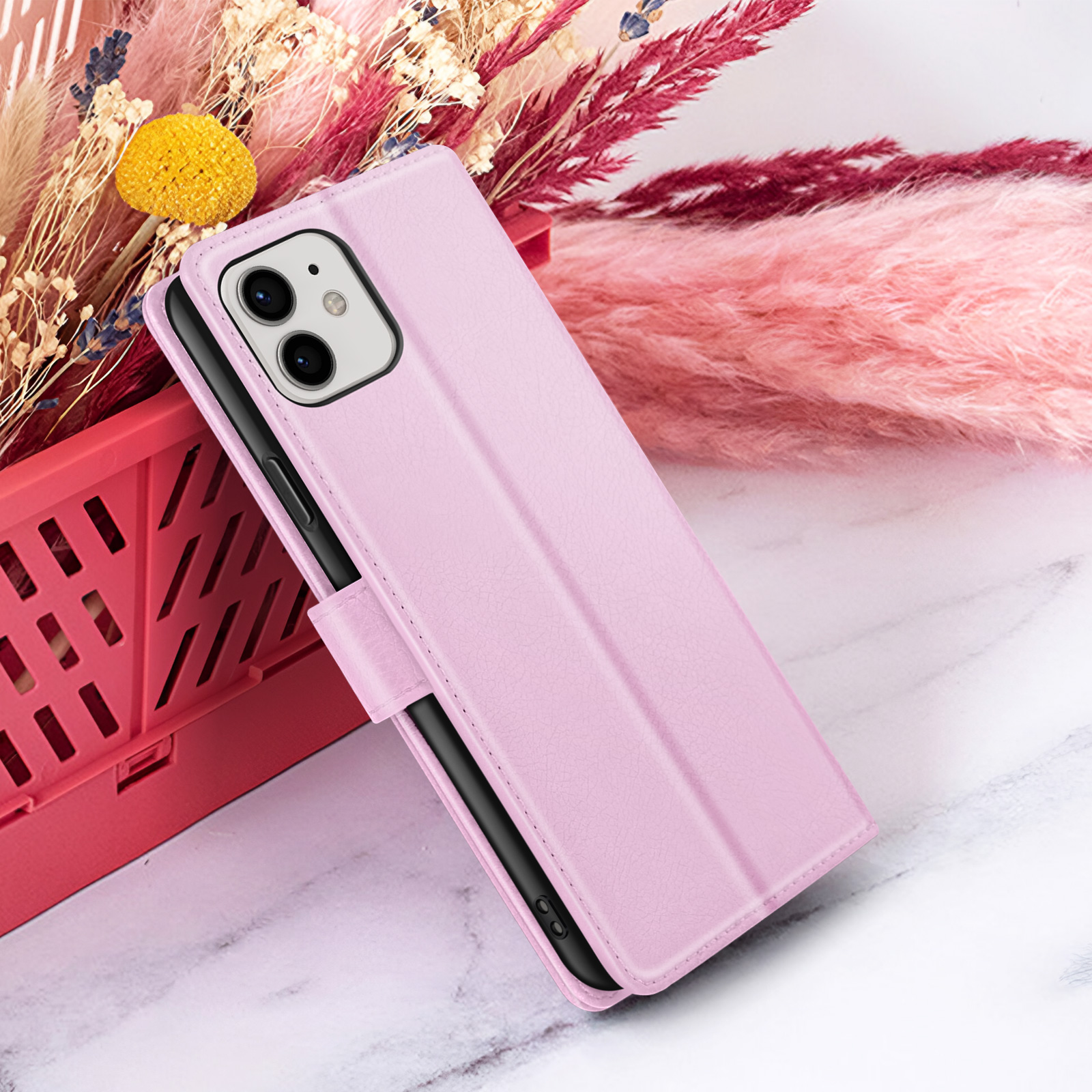 Een roze telefoonhoesje, open, met een telefoon erin. Het ligt in de buurt van roze en bruinrode decoratieve bloemen. Een gele bloem is zichtbaar.