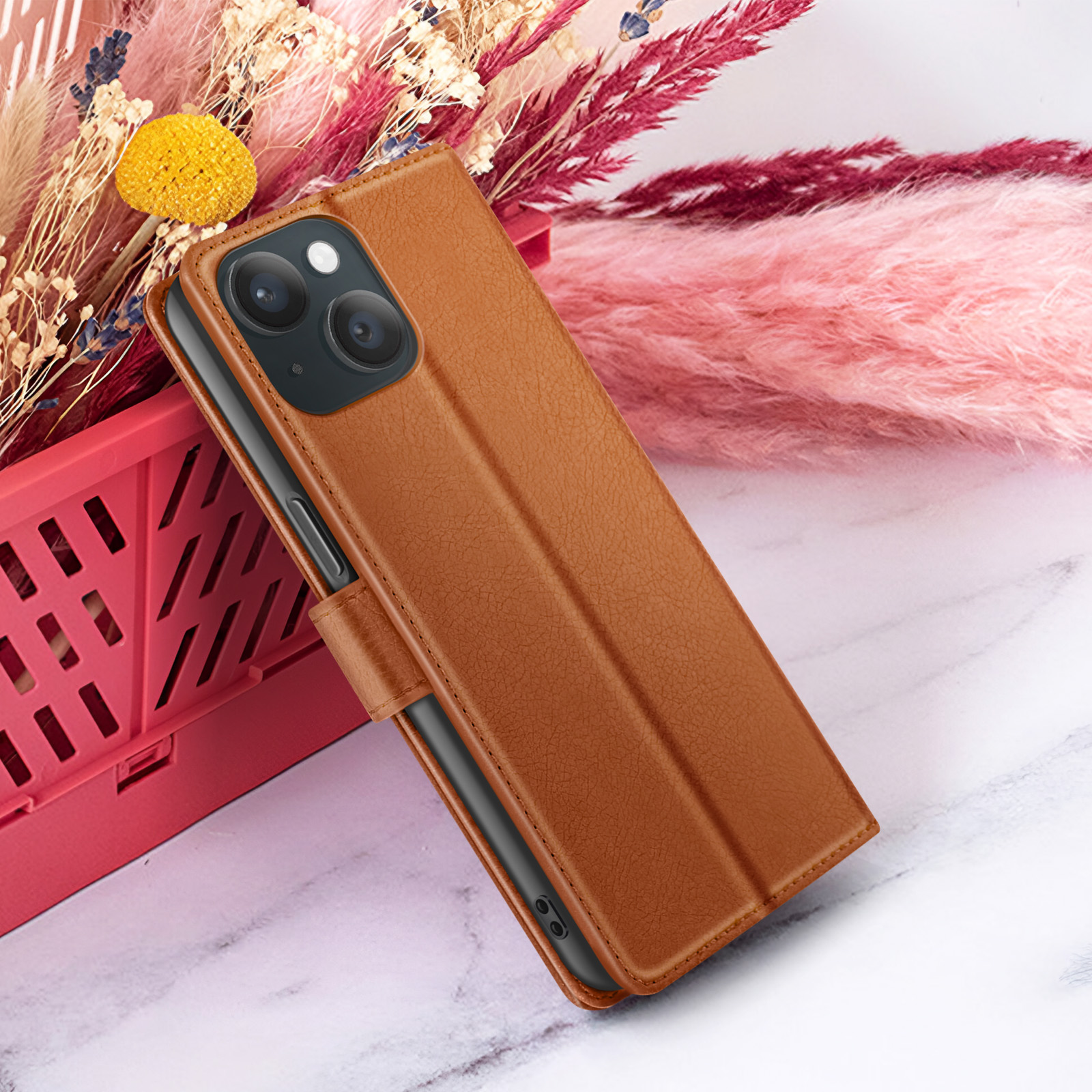 Un étui de téléphone en cuir marron est appuyé contre des fleurs dans un panier rouge sur une surface en marbre.
