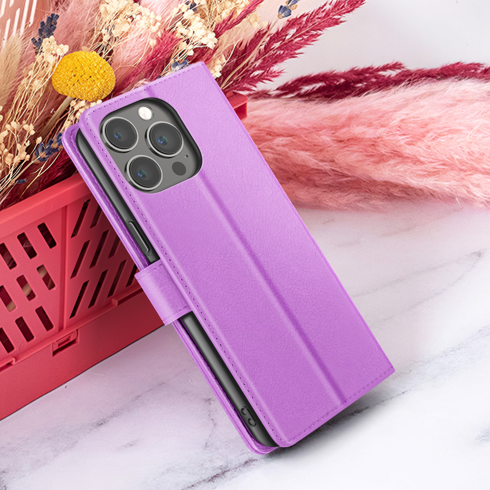 Un étui de téléphone violet est ouvert, révélant un téléphone avec trois caméras. Fleurs séchées et duvet rose en arrière-plan.